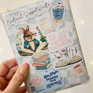 Puede incluir: Una funda de plástico transparente para un lector electrónico con varias pegatinas de colores. Las pegatinas presentan ilustraciones de libros, flores, un conejo, una taza de café y texto que dice "Emotional Support Kindle", "Read More Books", "Books are Magic", "Late Night Readers Club", "VIP Members Only", "Cozy Vibes".
