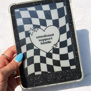 May include: A black glitter e-reader case featuring a wavy black and white checkered pattern on the screen. A white heart shape is centered on the display with the black text "emotional support kindle" inside, accompanied by two small star icons.