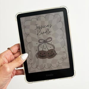 May include: A black Kindle e-reader with a clear protective case. The screen displays a design with the text "Jessica's Kindle" and two brown pumpkin illustrations. The background has a checkered pattern. The word "kindle" is at the bottom.