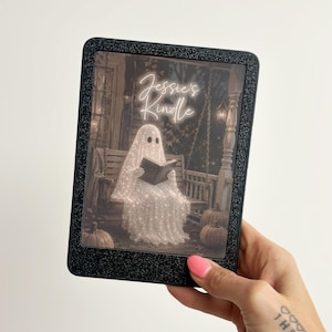 May include: A black glittery e-reader case featuring a screen display of a ghost sitting on a porch swing reading a book, surrounded by pumpkins. The text "Jessie's Kindle" is written in a white script font at the top of the screen display.
