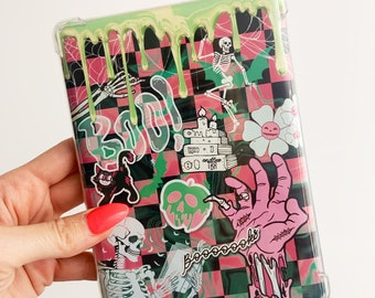 Op de afbeelding: Een doorzichtige tablethoes met een zwart-roze geblokte achtergrond en groen slijm dat van boven naar beneden druipt. De afbeeldingen bevatten skeletten, een zwarte kat, een stapel boeken, een pompoenbloem en een roze hand met de tekst Booooooks.