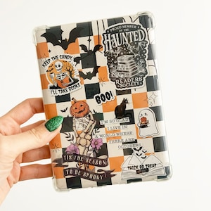 May include: A black and orange checkered case with Halloween-themed stickers. The stickers feature a skeleton holding a book, a pumpkin, a ghost, a cat, and a book with the text "Trick or Treat". The case also has a sticker that says "Proud Member of the Haunted Readers Society".