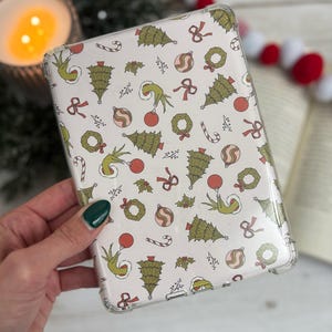 May include: A clear protective case for an e-reader with a festive pattern. The design features green Grinch hands, Christmas trees, ornaments, wreaths, and candy canes on a cream background. The case is held by a hand with a green painted nail.