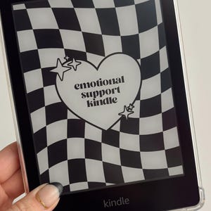 Może przedstawiać: Czytnik e-book&oacute;w Kindle z czarno-białym wzorem w szachownicę. Ekran wyświetla białe serce ze słowami "emotional support kindle" i akcentami gwiazd. Słowo "kindle" znajduje się na dole.