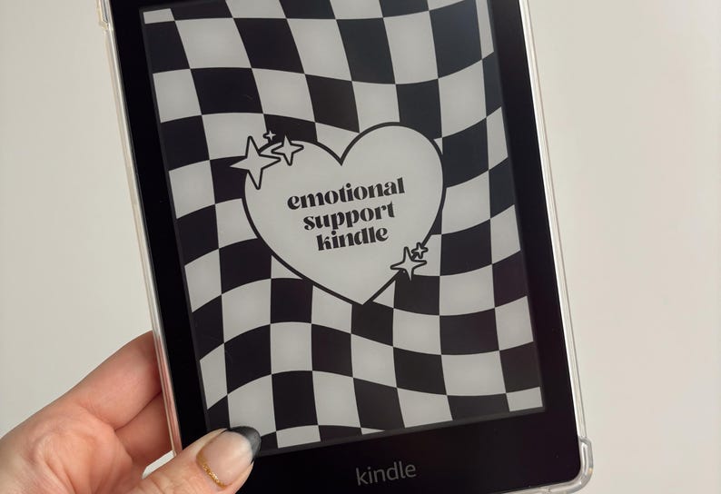 Może przedstawiać: Czytnik e-book&oacute;w Kindle z czarno-białym wzorem w szachownicę. Ekran wyświetla białe serce ze słowami "emotional support kindle" i akcentami gwiazd. Słowo "kindle" znajduje się na dole.