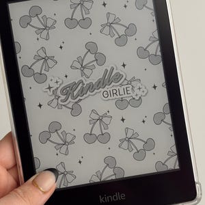 May include: A black and white Kindle e-reader with a clear case. The case has a sticker that says "Kindle Girlie" with a cherry pattern.