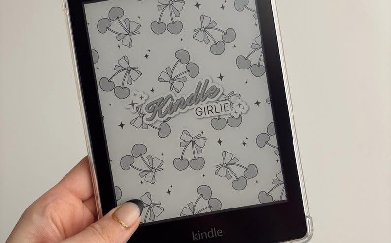 May include: A black and white Kindle e-reader with a clear case. The case has a sticker that says "Kindle Girlie" with a cherry pattern.