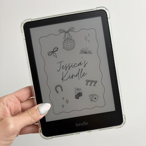 May include: A black Kindle e-reader in a clear case. The screen shows a decorative design with the text "Jessica's Kindle" and illustrations like a disco ball and playing cards. The e-reader is held in a hand.