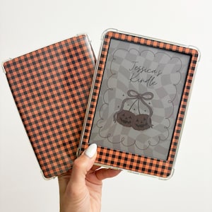 May include: A Kindle case and cover with a black and orange checkered pattern. The Kindle screen displays a gray and white checkered background with two pumpkin illustrations and the text "Jessica's Kindle".