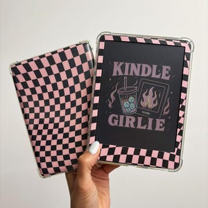 May include: A Kindle e-reader with a pink and black checkered case. The screen displays the text "KINDLE GIRLIE" with a graphic of a drink and flames. The case is clear with a pink and black checkered pattern.