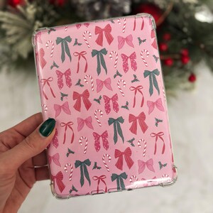 May include: Pink tablet case with a repeating pattern of bows, candy canes, and holly leaves. The bows are in shades of pink and green. The case has a clear, protective border.