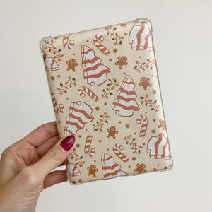 May include: A clear protective case featuring a festive Christmas pattern. The design includes gingerbread men, candy canes, Christmas trees, and string lights in red and white on a beige background. The case is held in a hand.