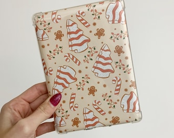 May include: A clear protective case featuring a festive Christmas pattern. The design includes gingerbread men, candy canes, Christmas trees, and string lights in red and white on a beige background. The case is held in a hand.