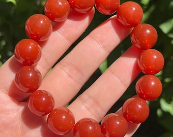 May include: A red-orange beaded bracelet. The bracelet is made of smooth, round beads. The beads are a rich, translucent color. The bracelet is held in a hand against a blurred green background.