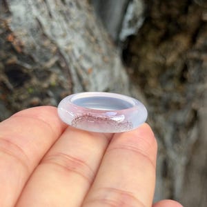 May include: A translucent ring with a light pink and gray hue. The ring has a smooth, rounded design and a subtle pattern of darker inclusions. The ring is a piece of jewelry.