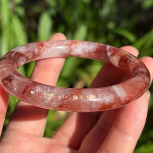 May include: A smooth, circular bangle bracelet crafted from translucent quartz featuring deep red and rust-colored mineral inclusions throughout the stone. The polished surface displays internal fractures and shimmering golden flecks.