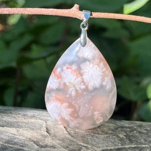 May include: A teardrop-shaped pendant with a silver bail. The pendant features a translucent stone with white and orange floral patterns. The stone is approximately 1.5 inches long and 1 inch wide. The pendant hangs from a small branch.