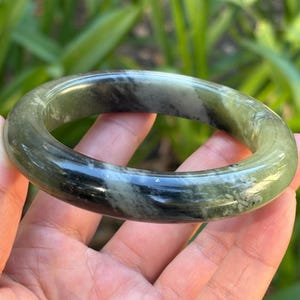 May include: A smooth, polished bangle bracelet crafted from mottled green and black stone. The circular jewelry piece features a rounded profile with a glossy finish, showcasing natural color variations and dark mineral inclusions throughout.