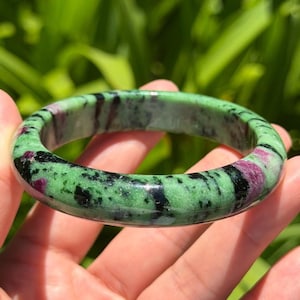 Puede incluir: Un brazalete rígido pulido hecho de piedra zoisita, con un color base verde vibrante y parches irregulares distintivos de color rojo magenta intenso, junto con vetas de inclusiones minerales negras en toda la superficie lisa y redondeada.