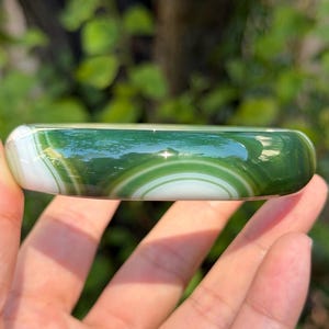 May include: A polished bangle bracelet featuring a smooth, rounded surface with alternating bands of deep forest green and opaque white. The stone displays natural concentric circular patterns and a glossy finish that reflects light across its curved exterior.