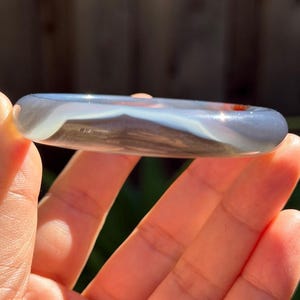 May include: A smooth, translucent bangle bracelet featuring a polished surface with swirling bands of light gray, white, and dark gray tones. The circular accessory has a glossy finish that catches the light, revealing internal patterns and subtle color variations within the stone-like material.