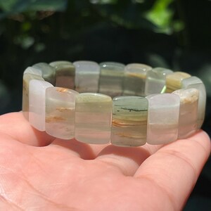 May include: A bracelet made of rectangular, translucent stones in shades of white, green, and brown. The stones are strung together to form a flexible band. The bracelet is held in a hand, with a blurred green background.