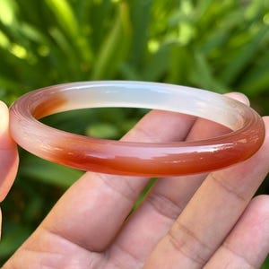 May include: A smooth, circular bangle bracelet crafted from translucent stone, featuring a gradient of colors transitioning from a deep, saturated reddish-orange on one side to a pale, milky white on the opposite side of the band.