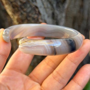 May include: A marbled gray and brown stone bangle bracelet. The bracelet has a smooth, polished surface with natural color variations. The design features a carved detail on one end. The bracelet is held in a hand.