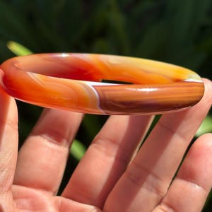 May include: A smooth, polished bangle bracelet crafted from banded agate stone. The circular jewelry piece features a gradient of warm colors, transitioning from deep reddish-orange and translucent amber to opaque brown, with distinct white mineral banding.