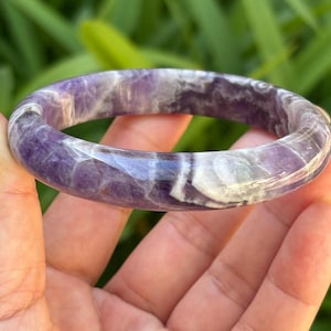 May include: A polished bangle bracelet crafted from natural amethyst stone, featuring a smooth, rounded surface with a variegated pattern of deep purple, lavender, and white mineral inclusions throughout the circular band.