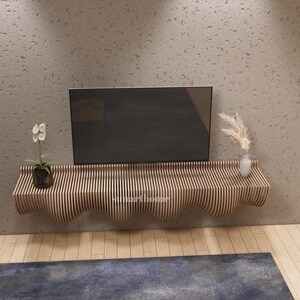 May include: A contemporary wooden floating shelf with a wavy design, supporting a flat-screen TV, a small plant, and a vase with decorative grass. The shelf is made of vertical wooden slats and is mounted on a textured grey wall.