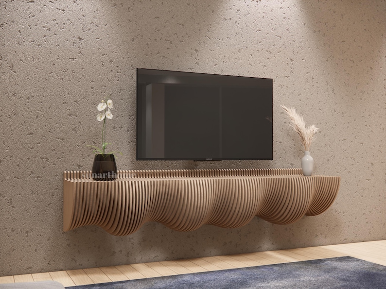 May include: A contemporary wooden TV unit with a distinctive, undulating design. The unit is crafted from light-toned wood and is fixed to a textured, light-brown wall. A black television is mounted above the unit. Decorative elements include a small plant and a vase.