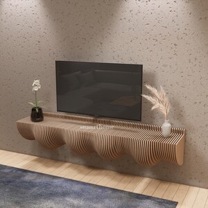 May include: A contemporary wooden TV unit with a distinctive, undulating form. The unit, constructed from vertical wooden slats, supports a flat-screen TV, a small black vase with a white flower, and a small white vase with decorative grass. The wall is a light grey colour.