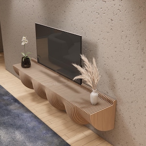 May include: A contemporary TV unit made of light-coloured wood, featuring a distinctive, undulating design. The unit is mounted on a textured wall and supports a large, black television. Decorative items include a vase with dried flowers and a small potted plant.