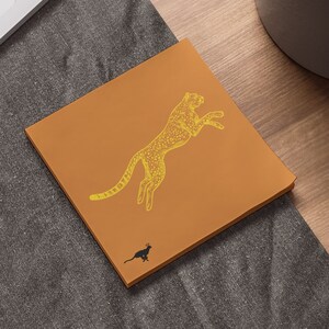 May include: A brown notepad with a yellow cheetah illustration and a small black cat illustration. The notepad is on a gray textured surface.