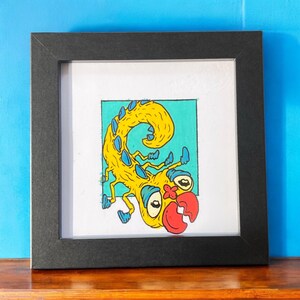 Könnte beinhalten: Gerahmtes Kunstwerk mit einer skurrilen gelben Kreatur mit blauen Akzenten und einem roten Mund. Das Kunstwerk ist vor einem türkisfarbenen Hintergrund in einem weißen Rahmen in einem schwarzen Rahmen angeordnet. Das Kunstwerk wird auf einer Holzoberfläche vor einer blauen Wand ausgestellt.