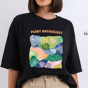Może przedstawiać: Czarny t-shirt z pomarańczowym napisem "PLANT ENTHUSIAST". Koszulka posiada kolorowy, abstrakcyjny wzór różnych roślin i liści. Słowo "black" jest napisane w prawym górnym rogu.