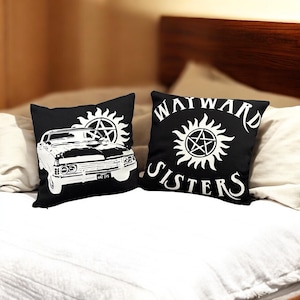 Può includere: Due cuscini neri con grafica bianca. Un cuscino presenta un design di auto classica, l'altro mostra il testo "WAYWARD SISTERS" con un simbolo di sole e pentagramma. I cuscini sono su un letto bianco.