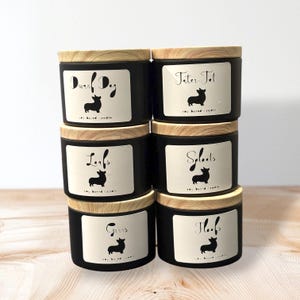 May include: Six black soy-based candles with wooden lids are stacked. Each candle features a white label with a black dog silhouette and a cursive name. The candles are displayed on a light-coloured wooden surface.