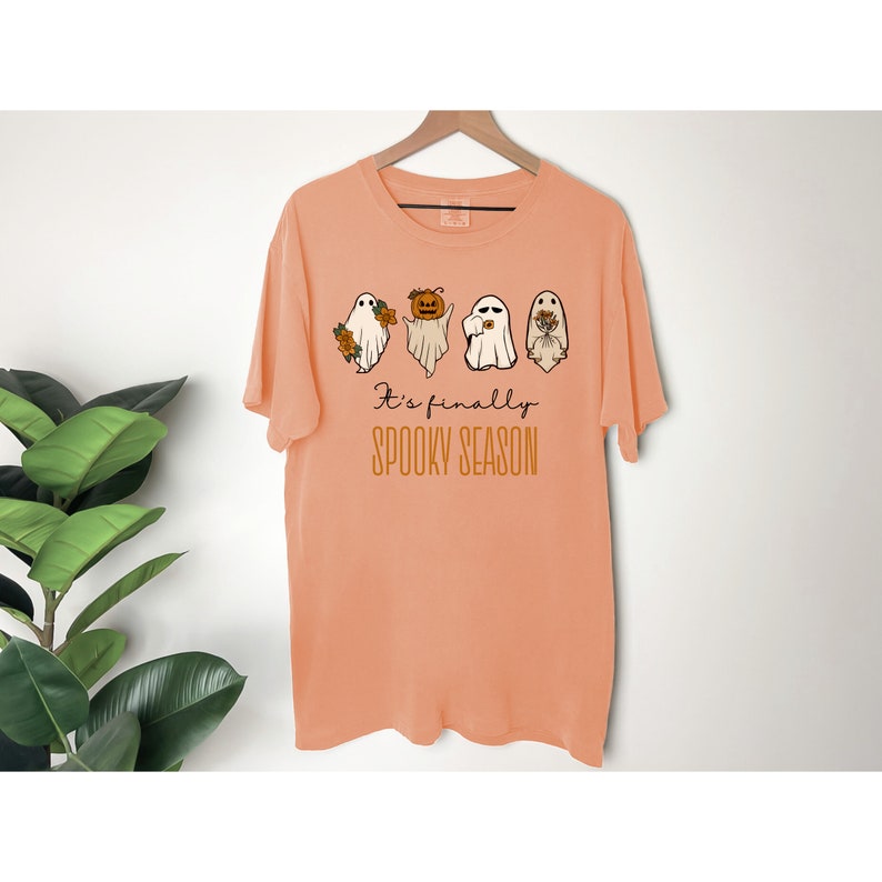 May include: Peach-colored t-shirt with a graphic of four ghosts. The ghosts are wearing different accessories, including flowers, a pumpkin, and a witch's hat. The text "It's finally spooky season" is printed below the ghosts.
