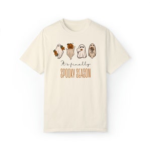 May include: A cream-colored t-shirt with a graphic of four ghosts. The ghosts are holding flowers, a pumpkin, a camera, and a bouquet of flowers. The text "It's finally spooky season" is printed below the ghosts.