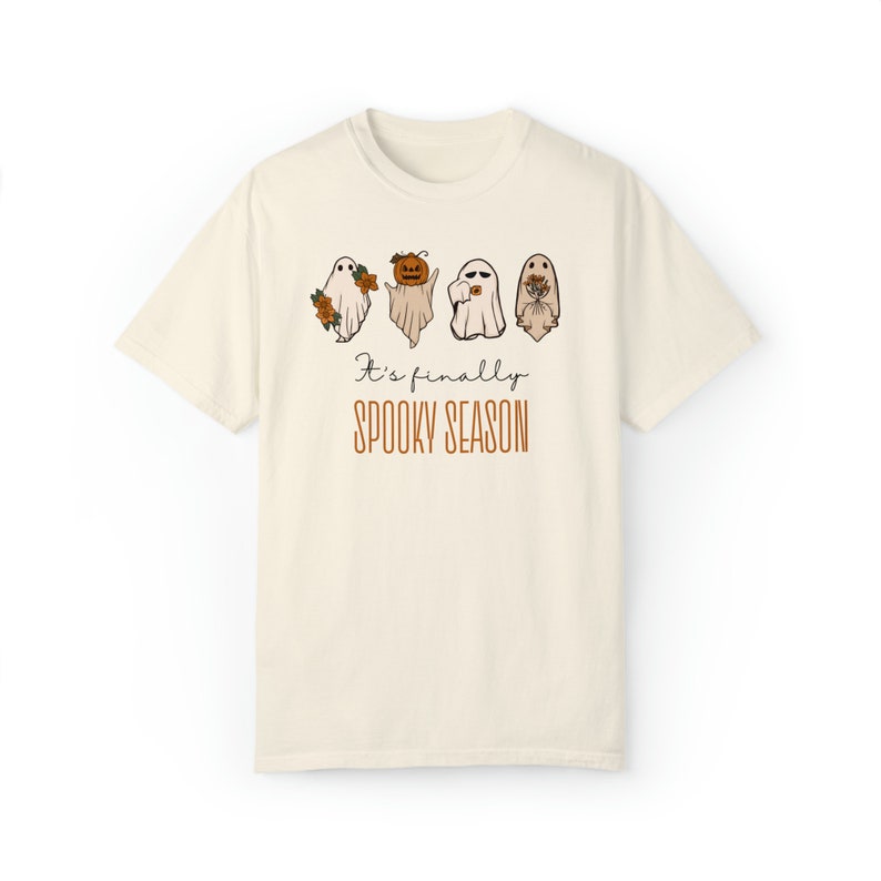 May include: A cream-colored t-shirt with a graphic of four ghosts. The ghosts are holding flowers, a pumpkin, a camera, and a bouquet of flowers. The text "It's finally spooky season" is printed below the ghosts.