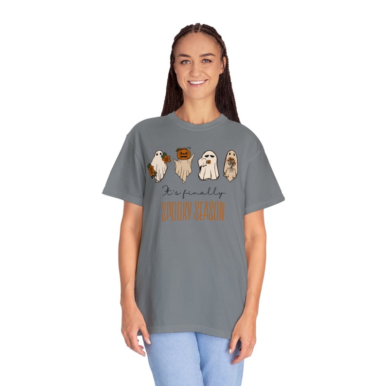 May include: A gray t-shirt with a graphic of four ghosts. The ghosts are holding a pumpkin, a cup of coffee, and a bouquet of flowers. The text "It's finally spooky season" is printed below the ghosts.