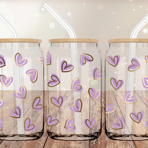 May include: Three clear glass drinking cups with bamboo lids and glass straws. Each cup is decorated with a pattern of lavender and gold hearts. The cups are displayed on a wooden surface.