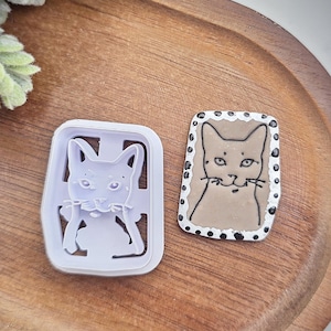 May include: Two cat-themed items sit on a wooden tray. One is a white plastic cat-shaped cookie cutter. The other is a hand-painted cat-shaped item with a brown cat and a dotted border. The tray has a light brown color.