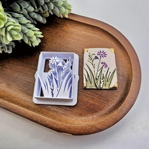 May include: A white plastic cookie cutter with a floral design next to a small, rectangular, decorated tile. The tile features a floral design with purple flowers and green grass. The items are on a wooden tray with a green plant in the background.