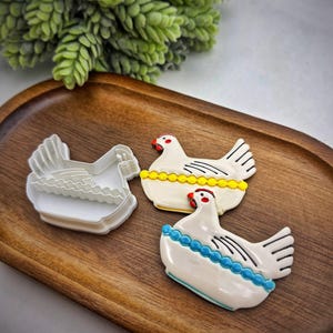 May include: Three chicken-shaped cookies on a wooden tray. Two cookies are decorated with white icing, black wing details, and red beaks. One cookie has a yellow beaded border, and the other has a blue beaded border. A white cookie cutter is also present.