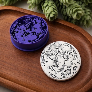 May include: A circular herb grinder featuring a vibrant purple interior with sharp, jagged teeth and a separate silver-toned lid embossed with a detailed, textured lunar surface pattern, both resting on a smooth, oval-shaped wooden tray.