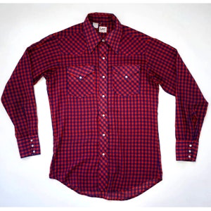 Puede incluir: Una camisa de manga larga de cuadros rojos y azules con cuello puntiagudo, dos bolsillos en el pecho y cierre de botones. La camisa tiene una etiqueta de la marca Lee.