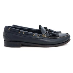 May include: A pair of navy blue leather loafers with tassel details. The shoes feature a classic design with gold-tone eyelets and stitching around the sole. The loafers are designed for comfort and style.
